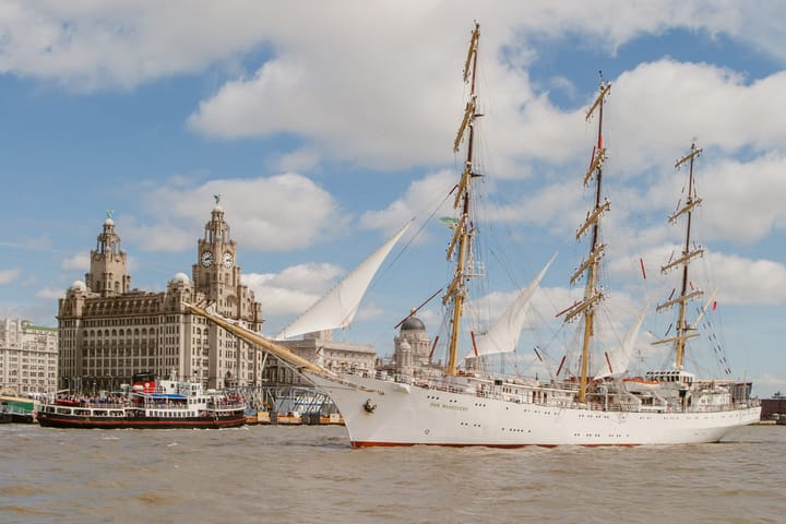A large tall ship sails past the Liverpool waterfront on a sunny day.