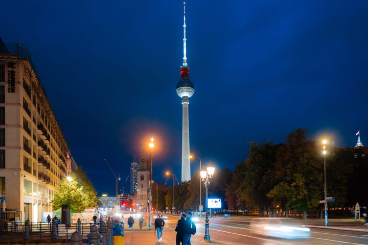 The illuminated TV Tower in Berlin at dusk, with pedestrians and traffic on the street.