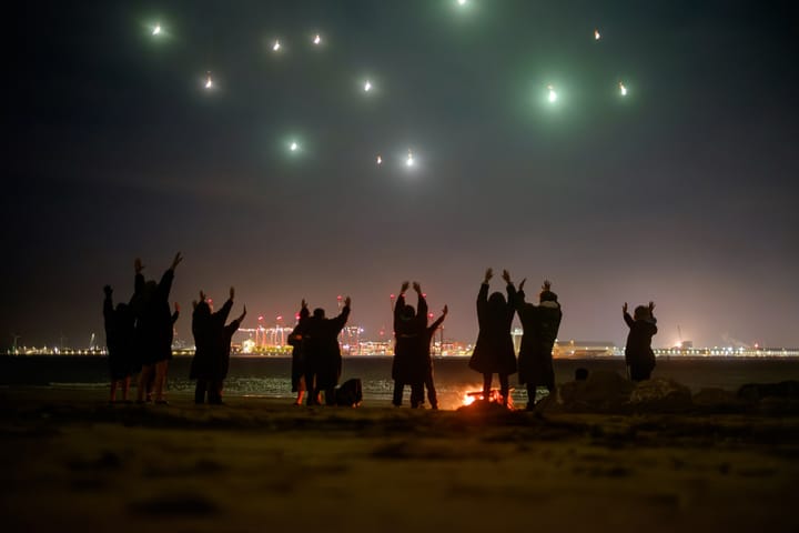 A group of people reaching to the sky at night while a firework explodes over head.