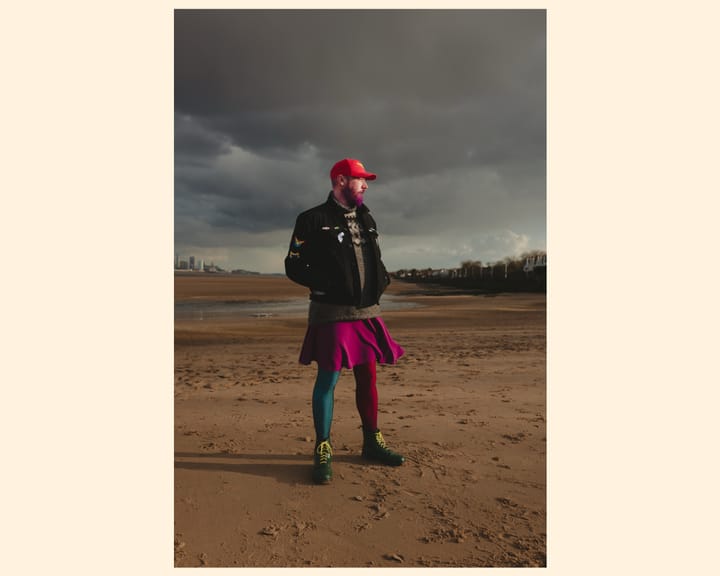 A person stands on a sandy beach under a dramatic sky, wearing a black jacket. They sport a vibrant purple skirt and colorful tights, complemented by bold green shoes.