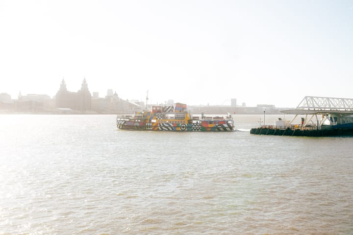 A colorful ferry boat sails on the River Mersey, with the Liverpool skyline in the background.