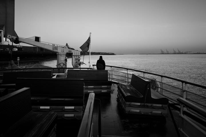 Black and white photo. A person stands alone on the upper deck of a ferry as it departs.
