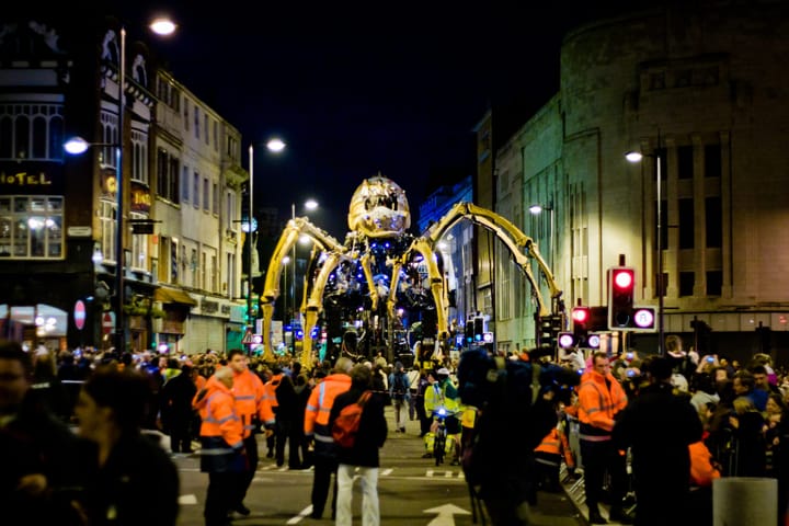 A giant, building size, mechanical spider walks down a busy street at night.