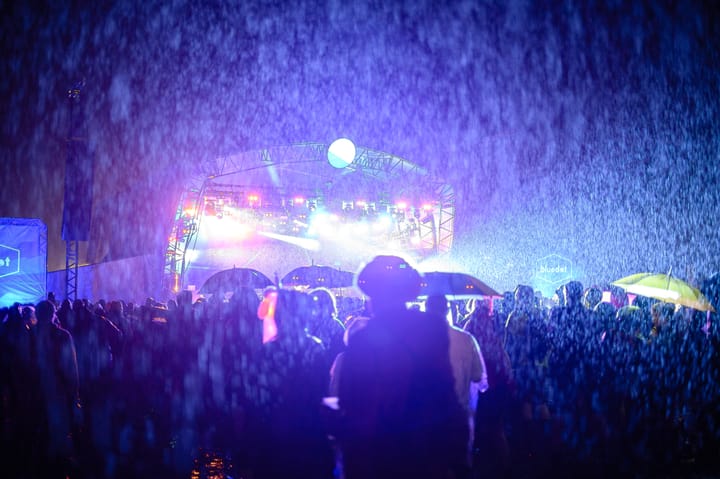 Rain backlit from stage lighting fills the sky as people watch a gig.