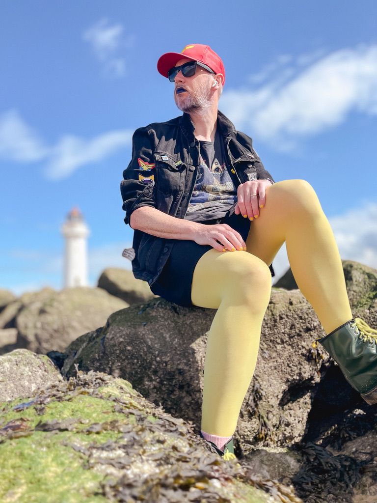 Non-binary person sat on rocks wearing yellow tights, black skirt, black denim jacket, black t-shirt with a lighthouse behind them.