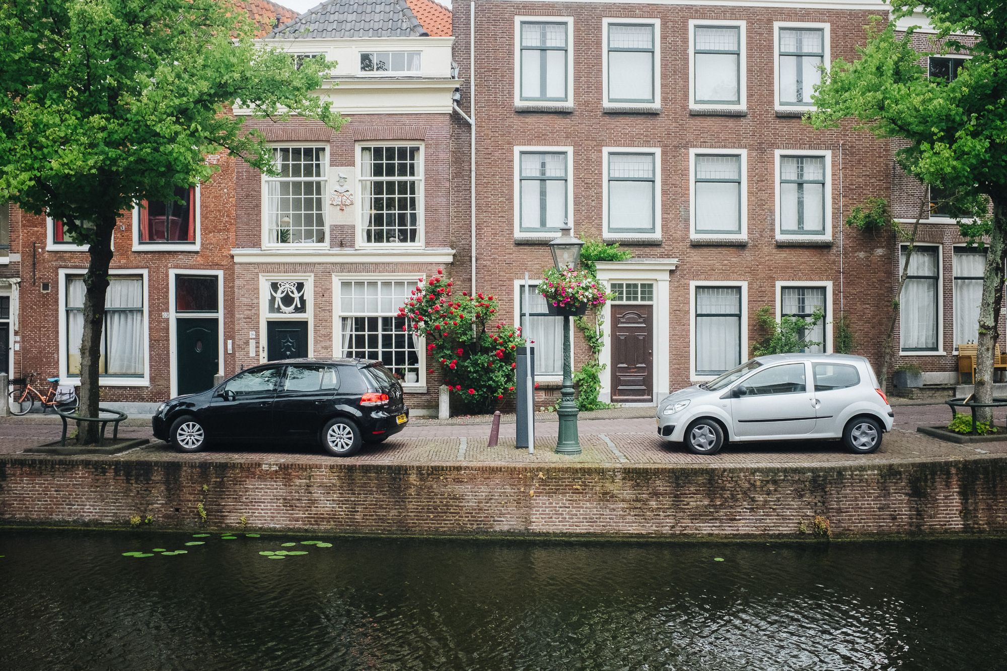 2 parked cars outside homes in Leiden. The canal is empty and there are big green trees either side of the houses.
