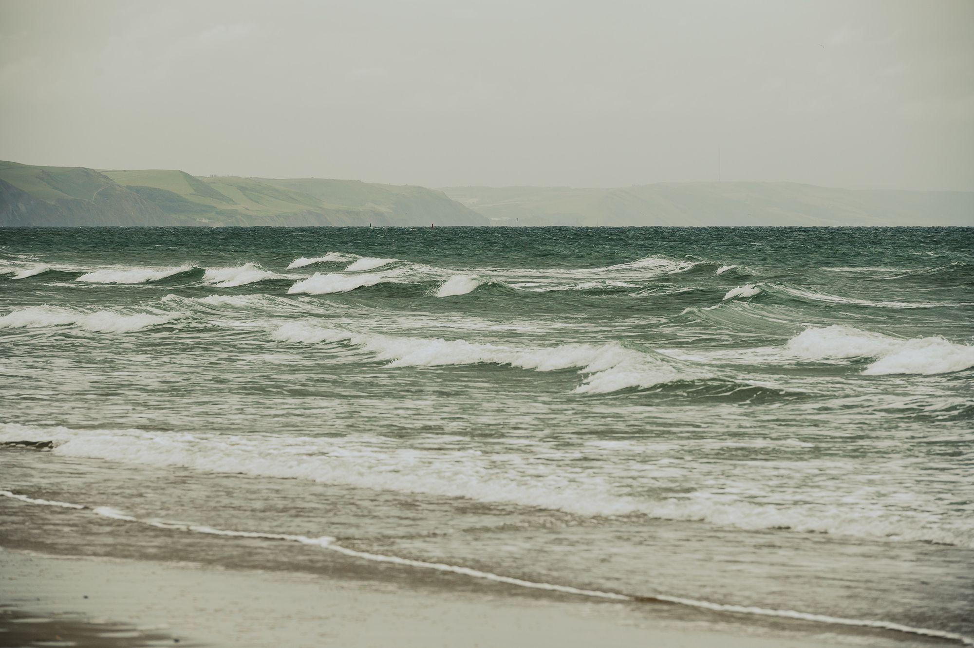 Large waves crashing onto a beach on an overcast day with big hills on the horizon.