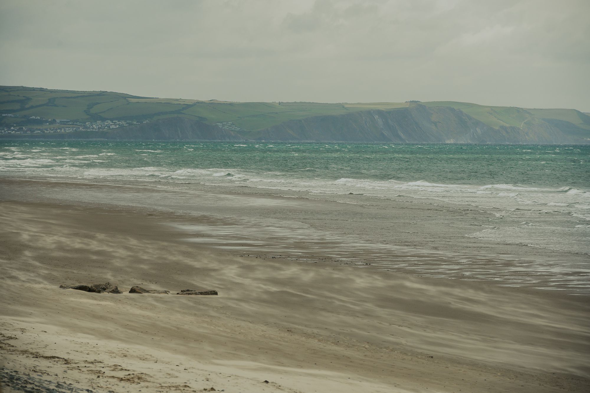 On a windy day sand flows across a beach while the tide goes out. In the distance there are cliffs and big hills.