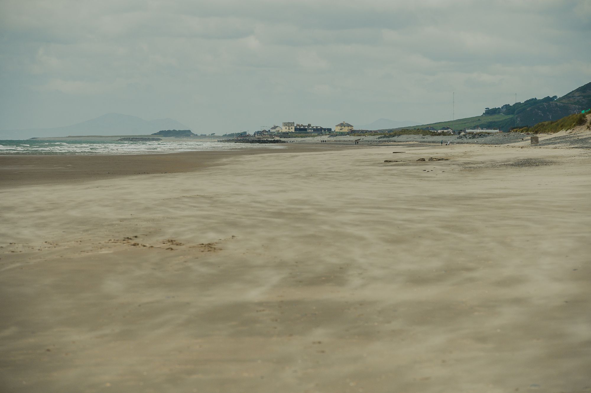 The wind is blowing sand along the beach on a cloudy day. There are buildings on the horizon and a mountain behind.