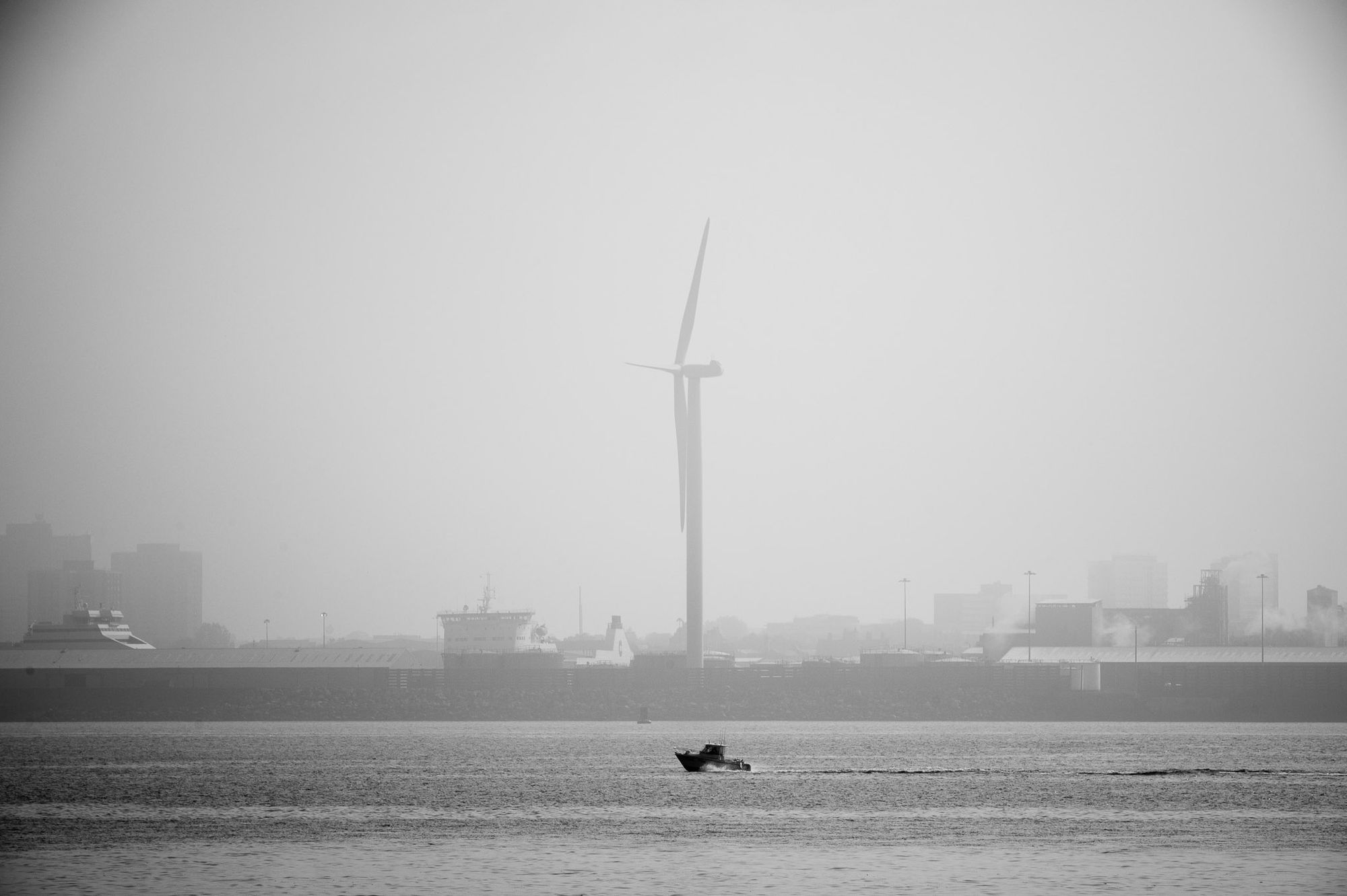 A small boat cruises along the River Mersey. The docks and a large wind turbine are in the background.