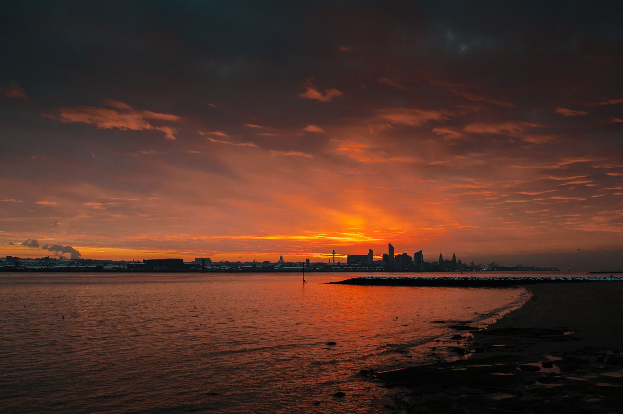 Red sky over the city of Liverpool. The tide is half in and there is snow on the rocks.