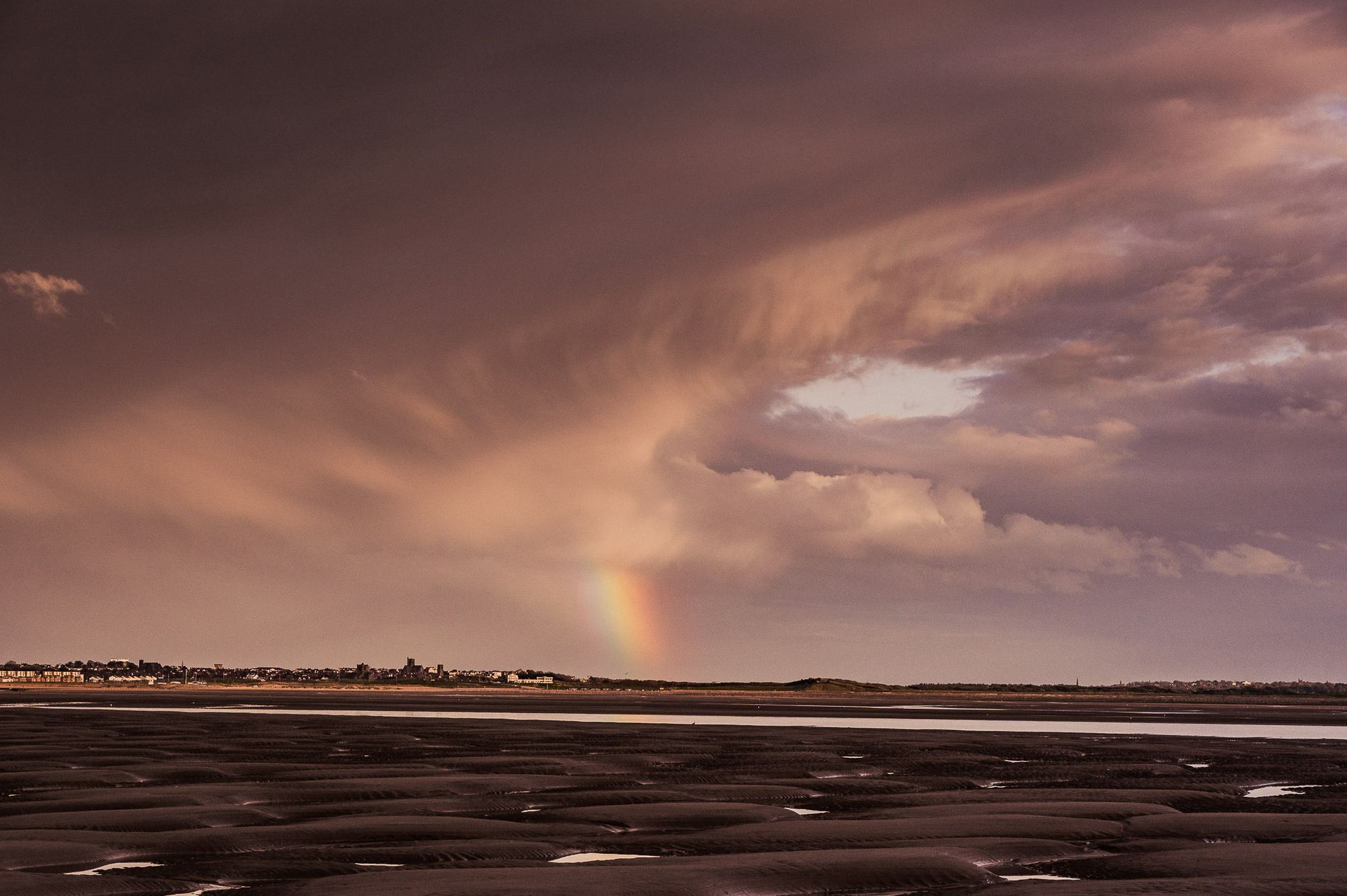 A partial rainbow sits in front of a large cloud over a beach