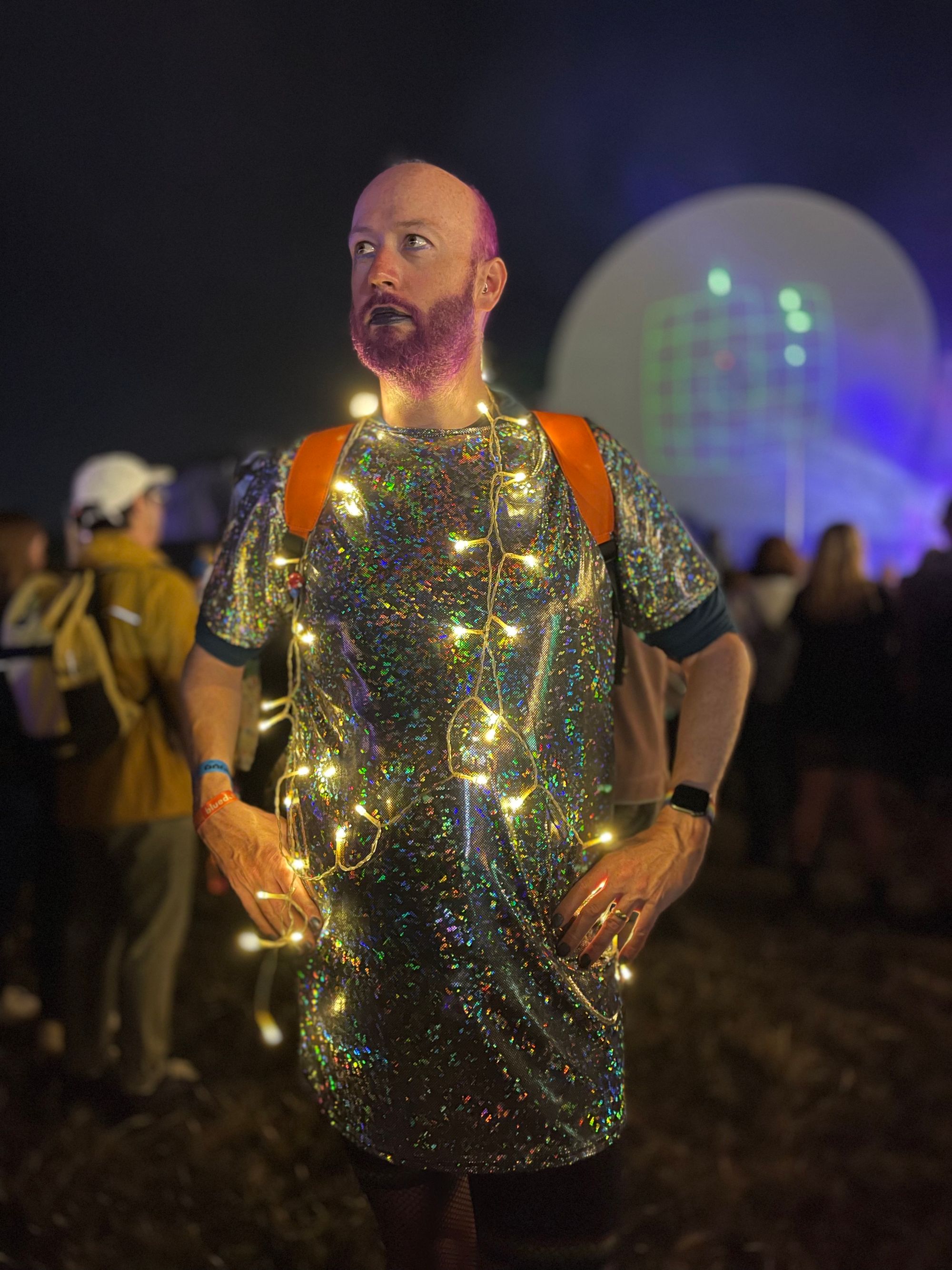 Non-binary person in sparkly dress wearing led lights standing in front of a radio telescope.
