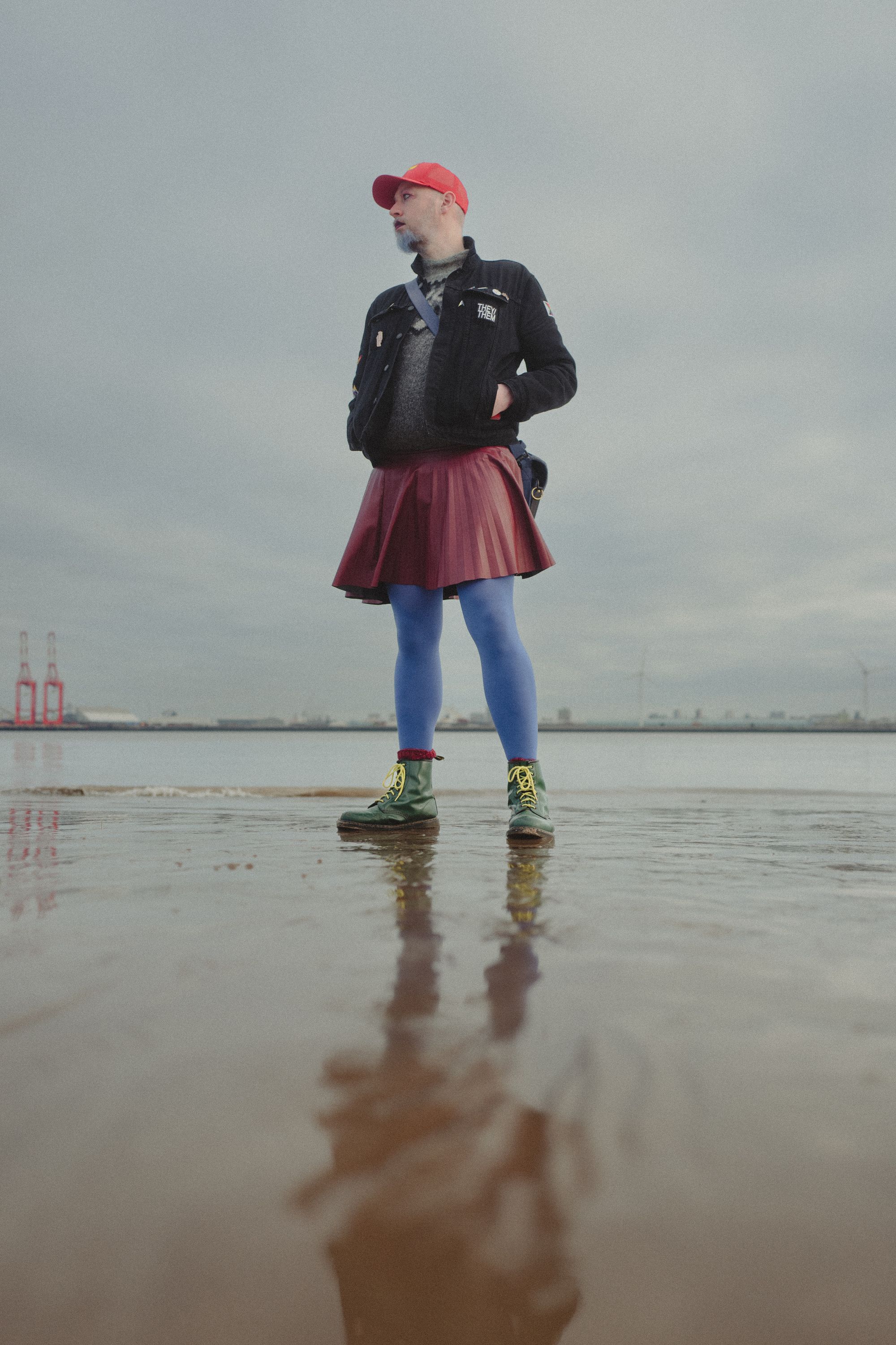 Non-binary person wearing black denim jacket, green boots, red skirt, blue tights and a red cap standing in front of a river.