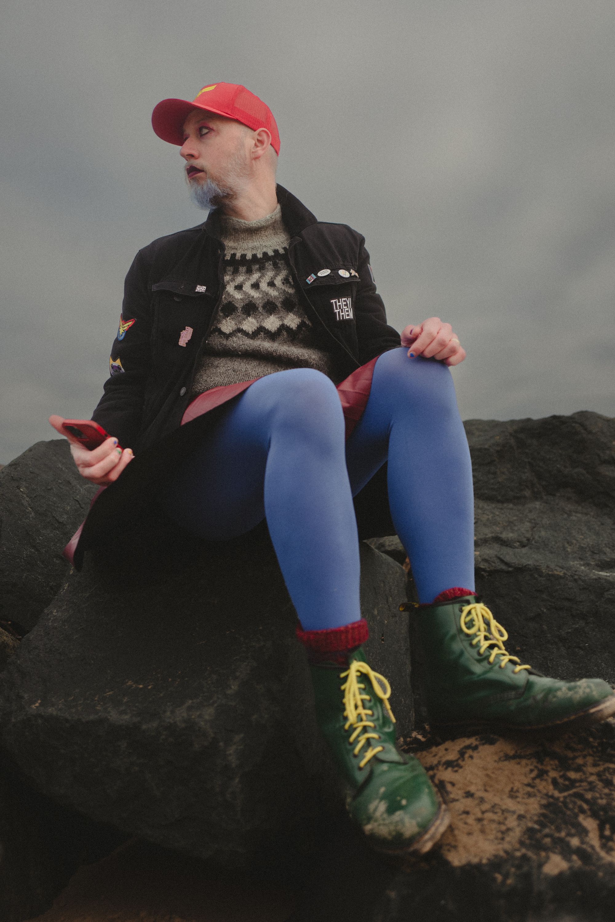 Non-binary person wearing black denim jacket, green boots, red skirt, blue tights and a red cap sat on rocks.