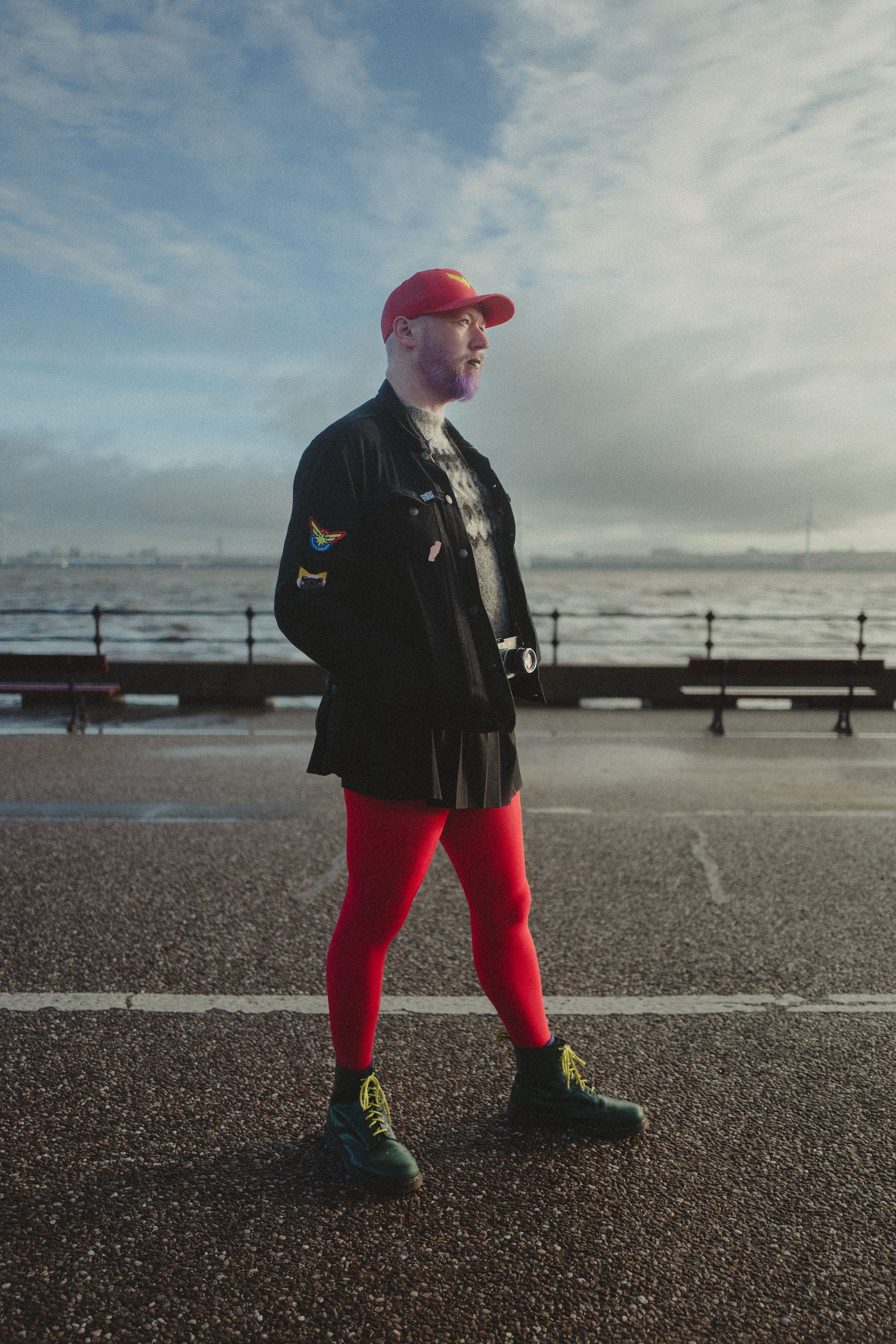 Non-binary person wearing black denim jacket, green boots, black skirt, red tights and a red cap standing on a promenade at high tide.