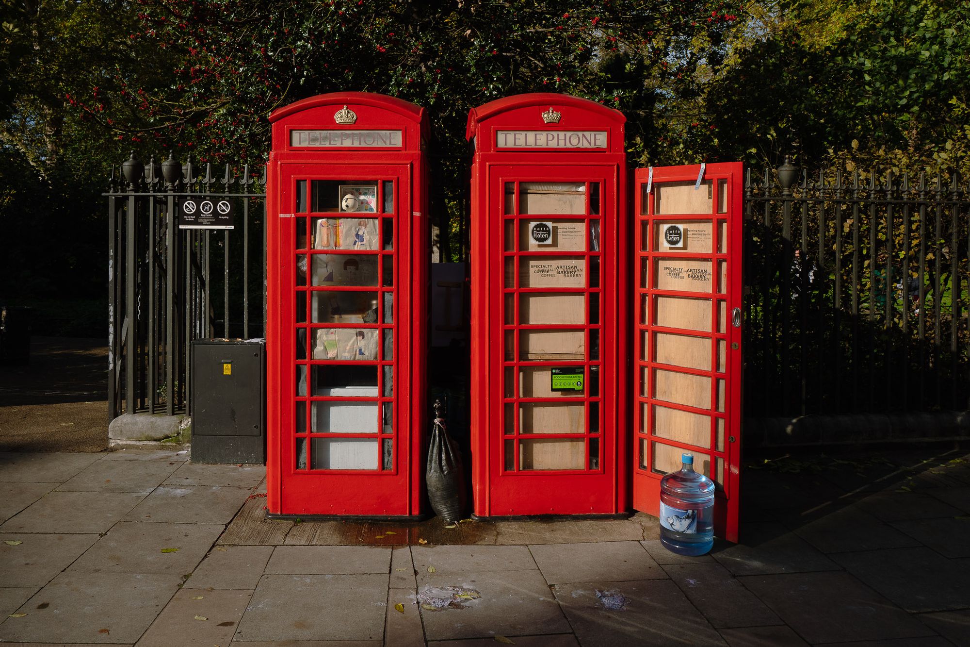 Two red telephone boxes are nicely lit in the evening sun outside a park. One is a small coffee shop.