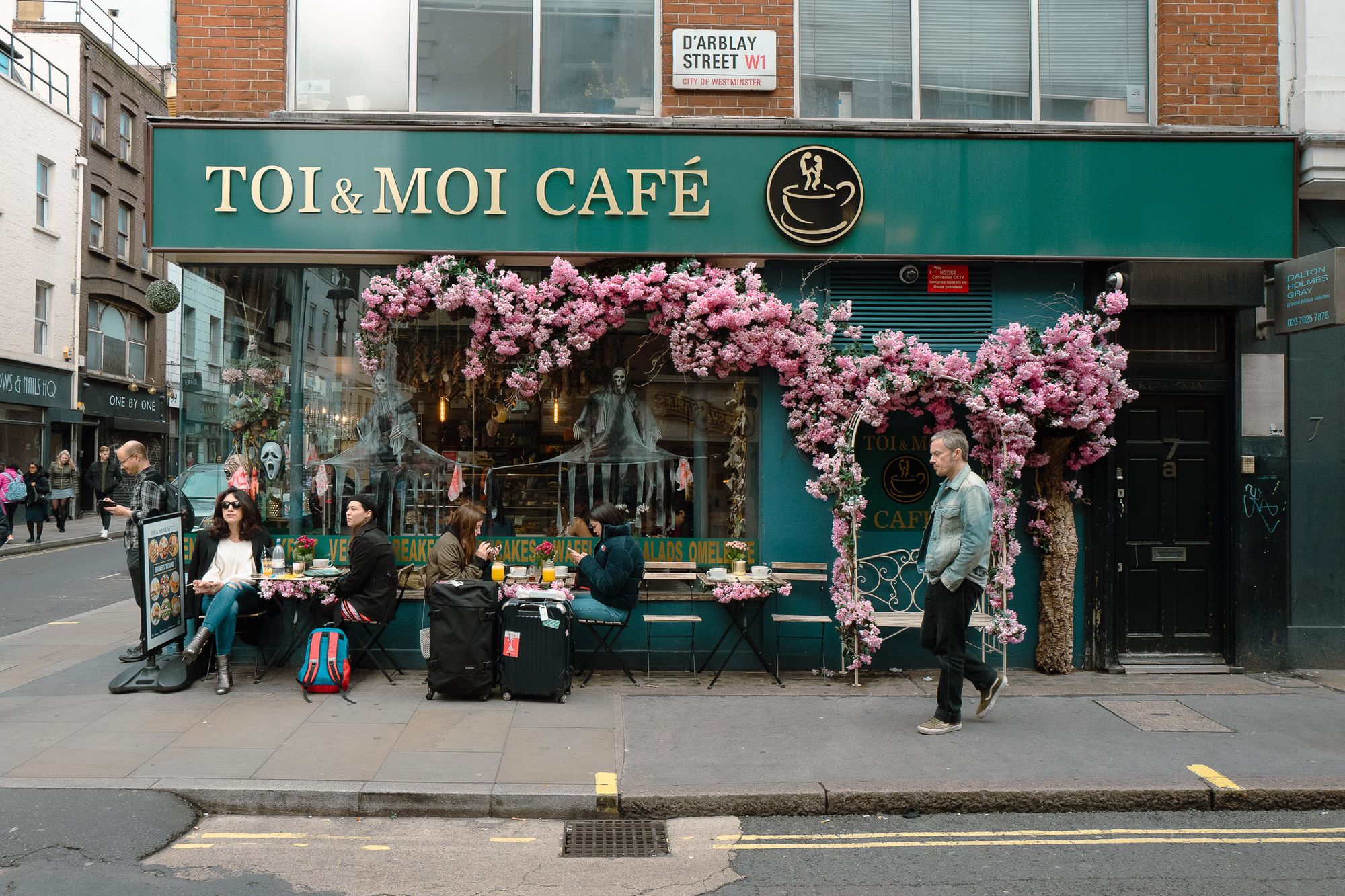 Toi & Moi Cafe in London. There are people sat outside. The cafe is painted green and there are pink flowers flowing around it. 