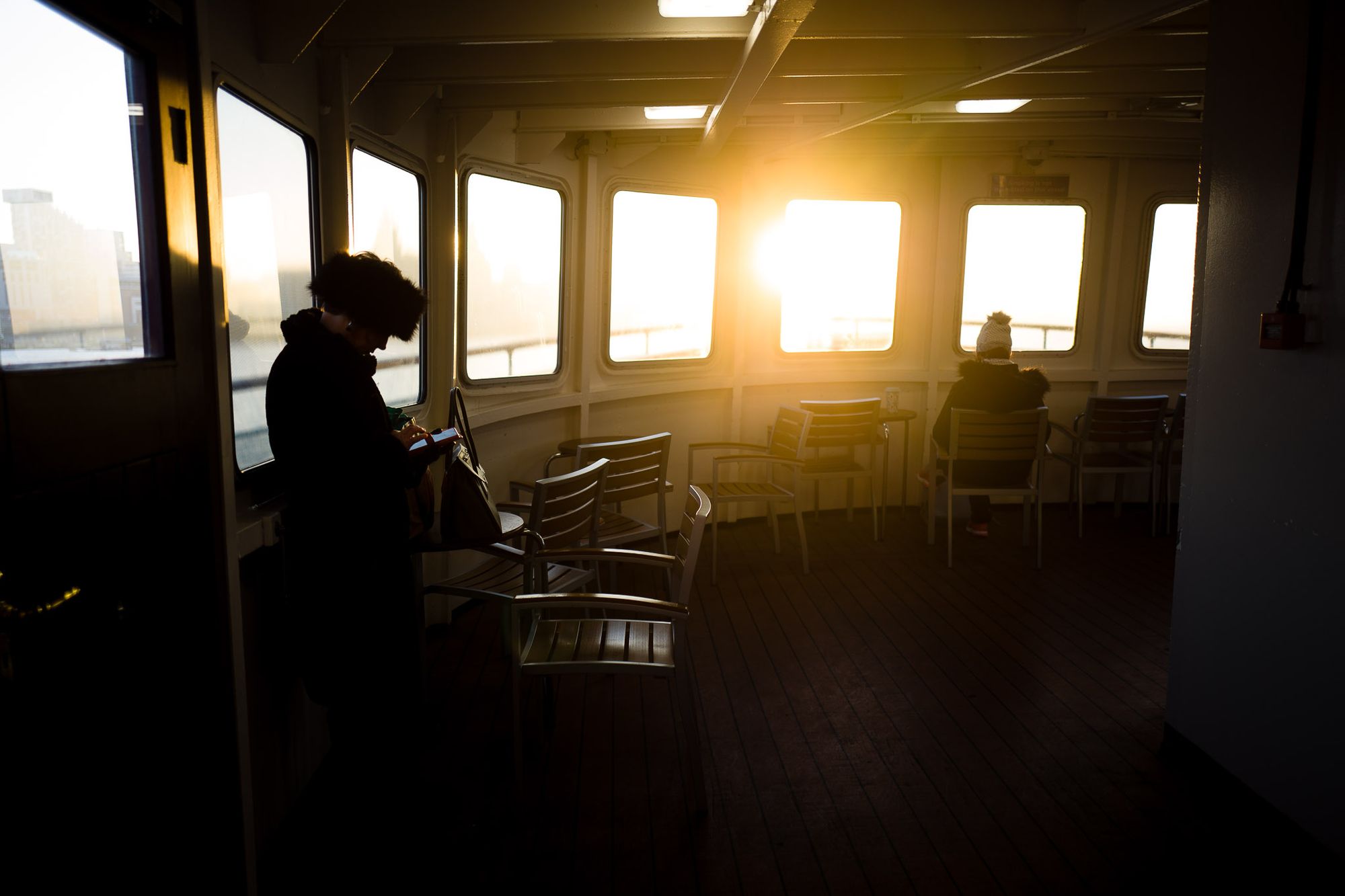 Sunrise through the window of a Mersey Ferry. A person stands on the left and another sits on the right.