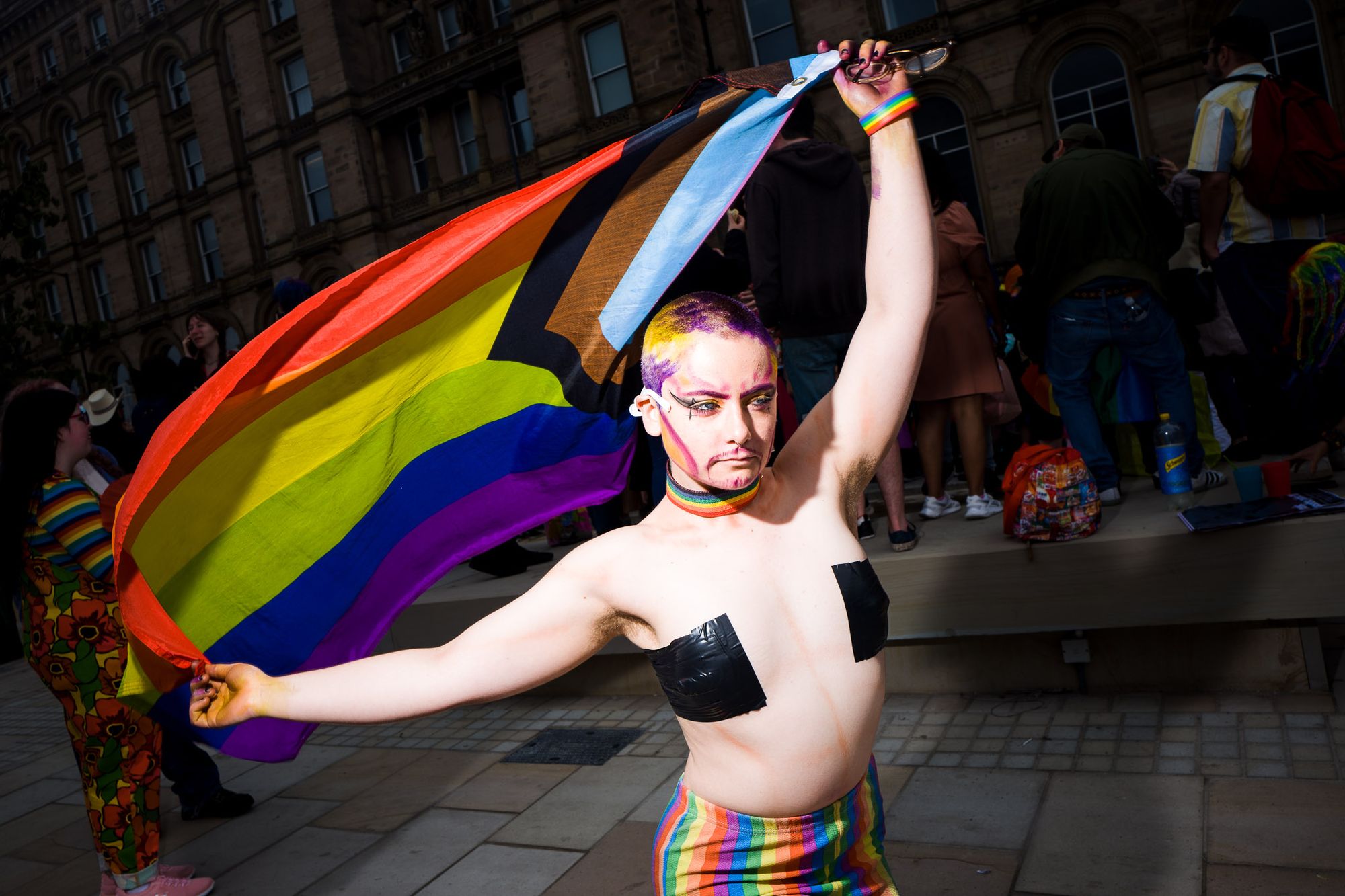 A person wearing black tape over their chest waves the progress pride flag around.