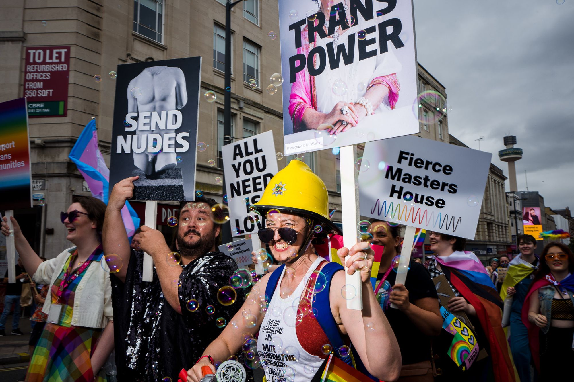 People from National Museums Liverpool march with placards. One says "Fierce Masters House". Another says "All you need is love." Another says "Trans Power" with a photo of April Ashley on. The last says "Send Nudes" over a photo of a marble statue.