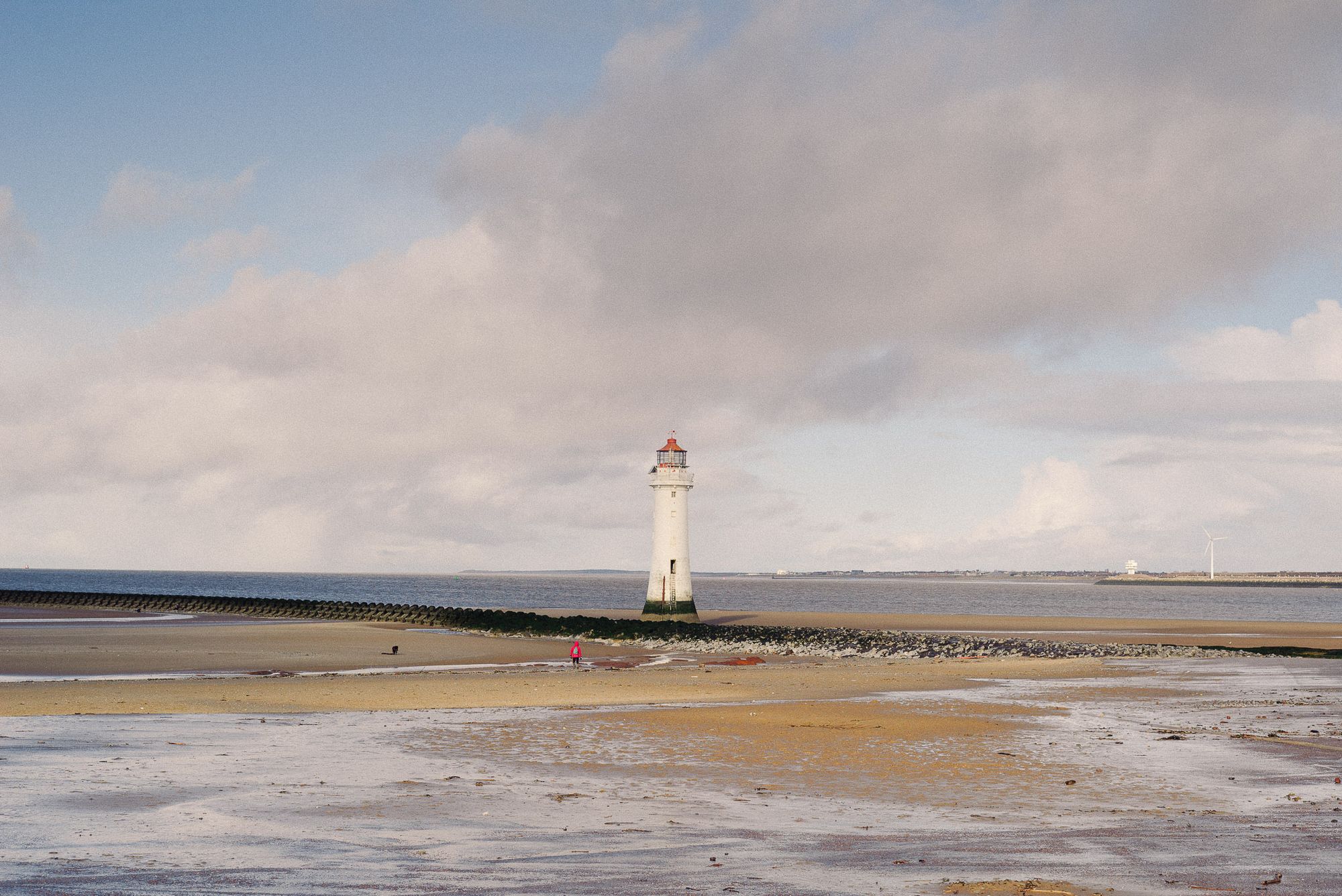 New Brighton lighthouse on a mostly clear day. The tide is out and people are walking on the beach.