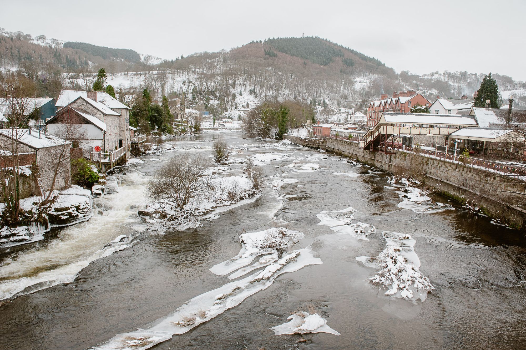 A river runs through the town of Llangollen. On the right is the train station. On the left are houses. Snow is on the rocks in the river and the banks.