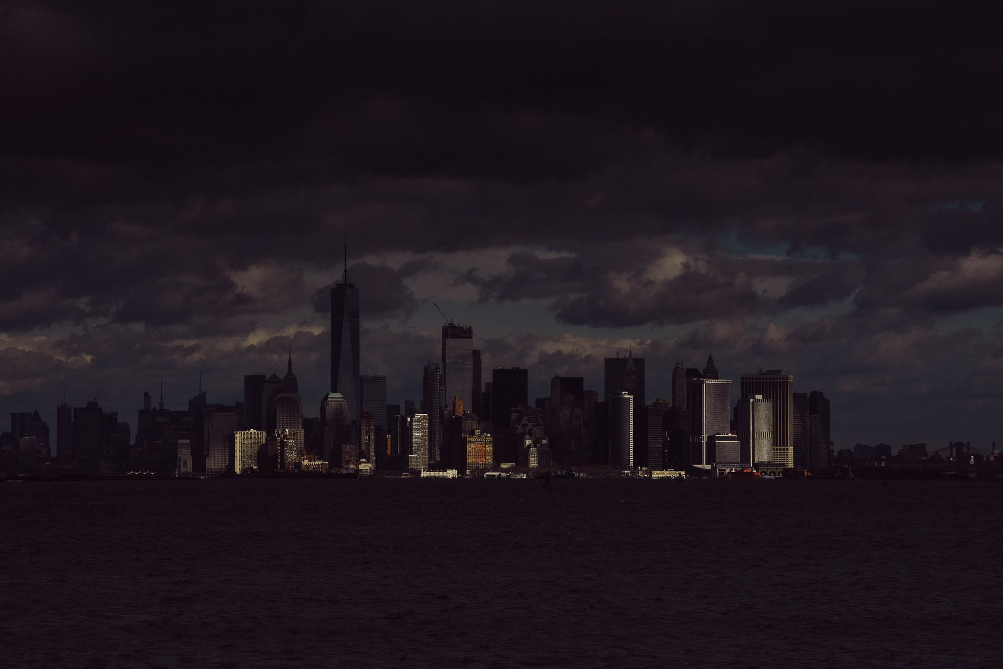 A dark and moody photo of the island of Manhattan. Just a few buildings are lit by sunshine.