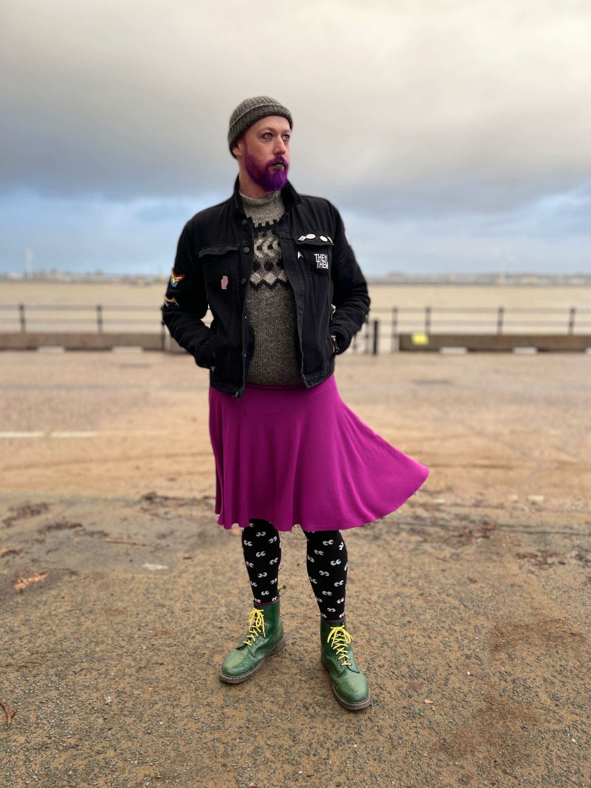 Non-binary person wearing green boots, tights with eyes on, purple skirt, Icelandic jumper and coat.