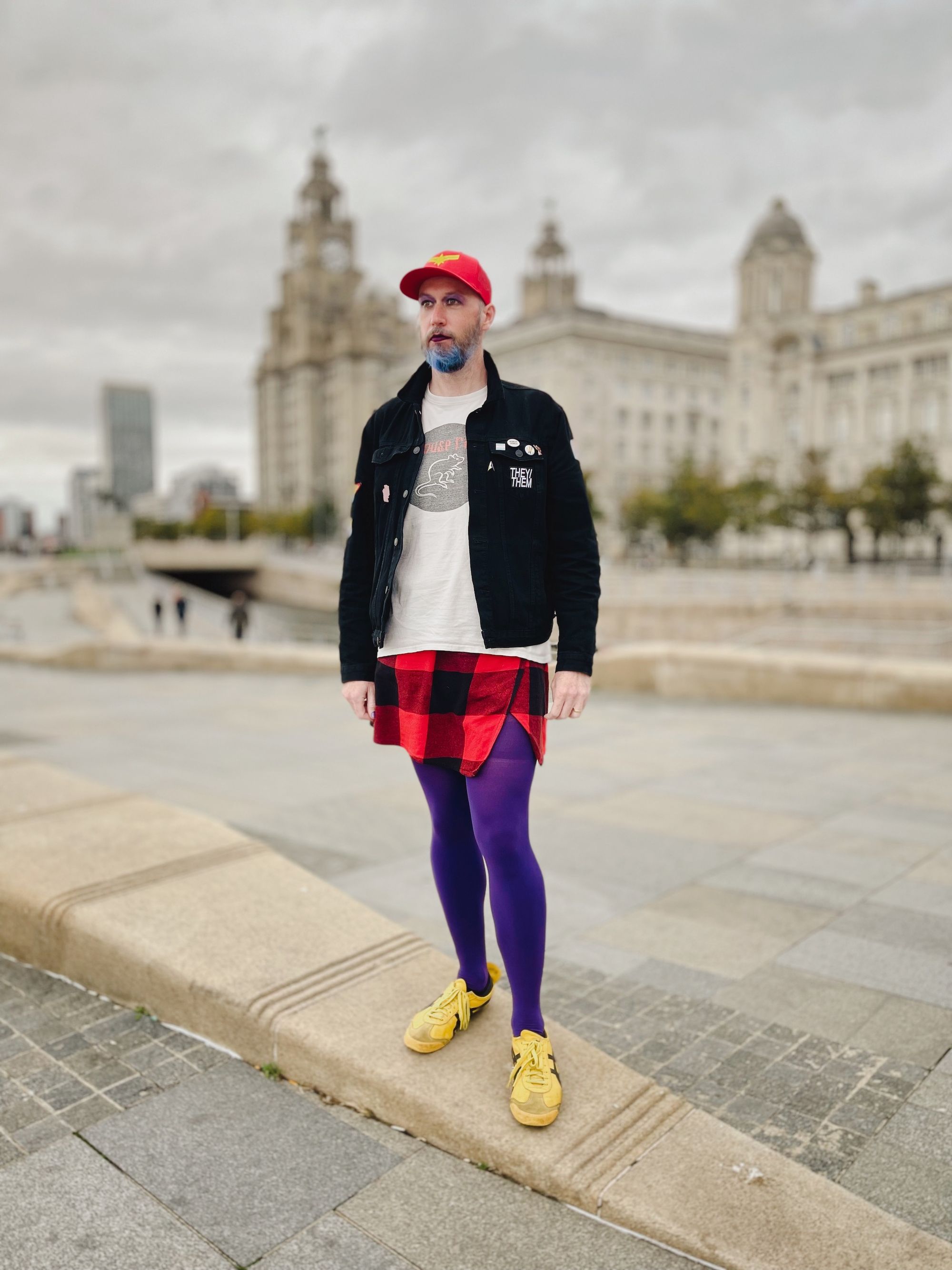 Non-binary person wearing purple tights, yellow shoes, a red check skirt, white t-shirt and black coat outside Liverpool waterfront.