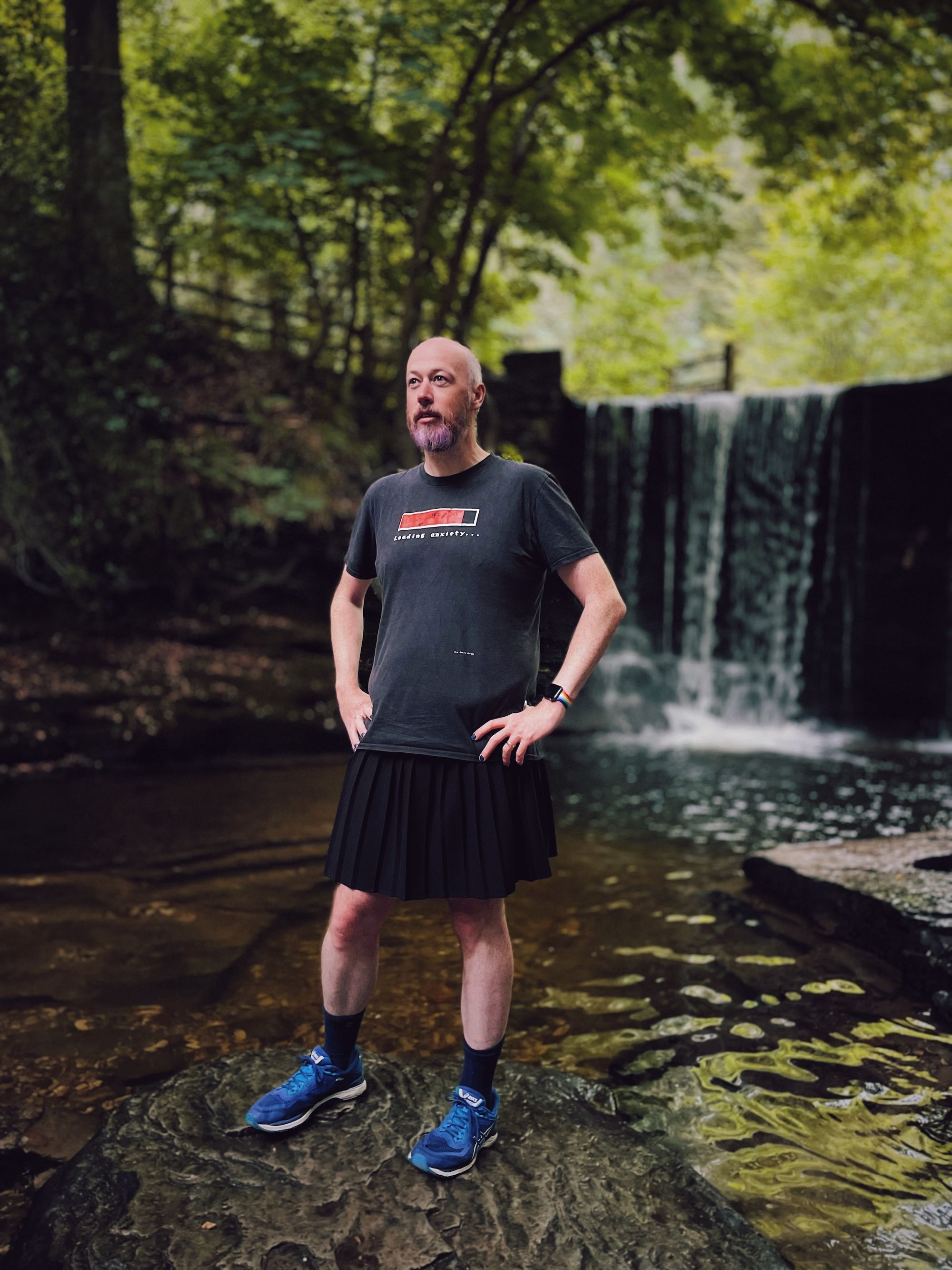 Non-binary person wearing a black skirt and t-shirt standing on rocks by a waterfall.