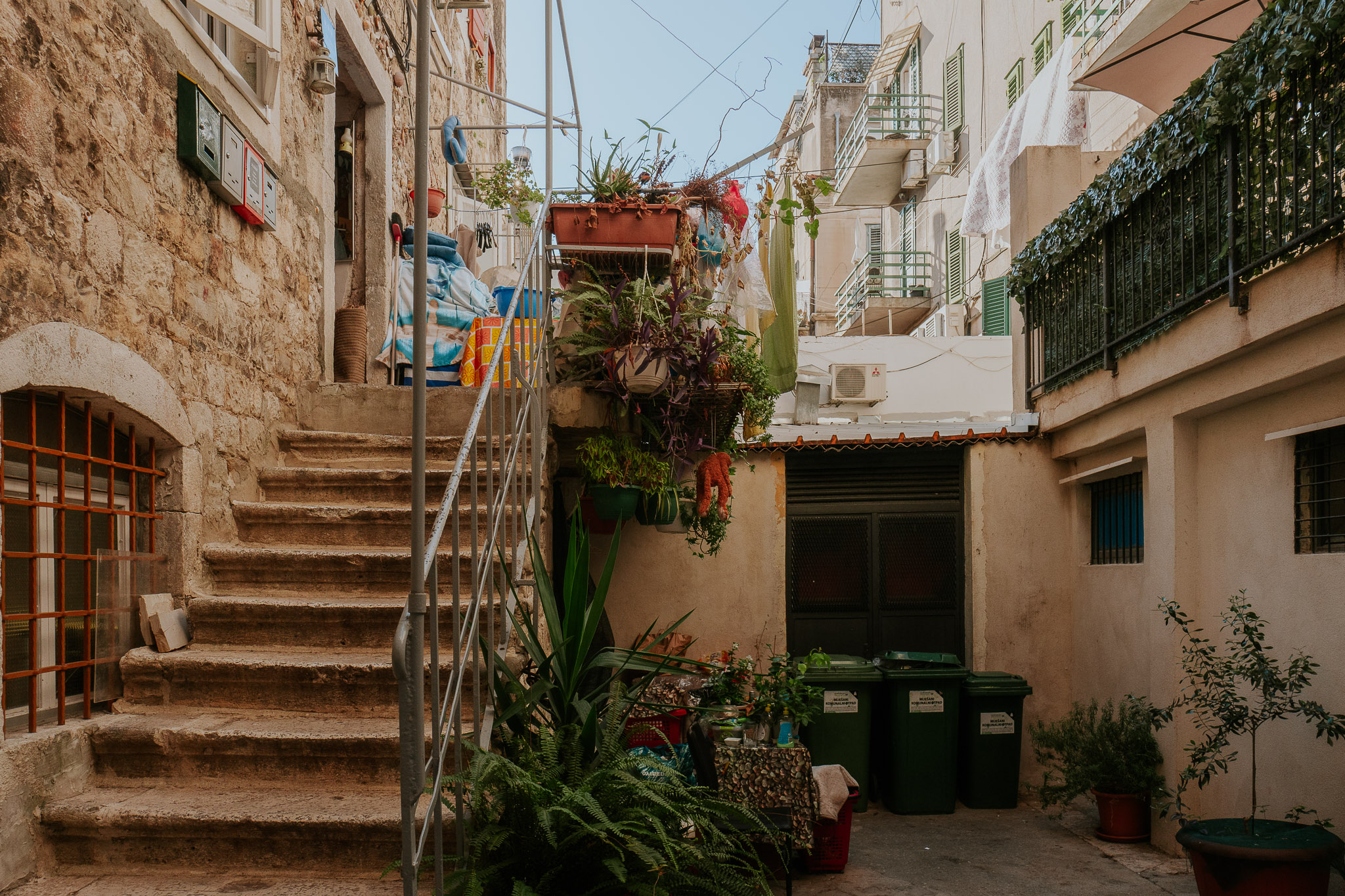 The backs of old houses in Split, Croatia. There are stairs on the left and a courtyard on the right. In the courtyard are plants and green bins. There are lots of balconies above with green railings.