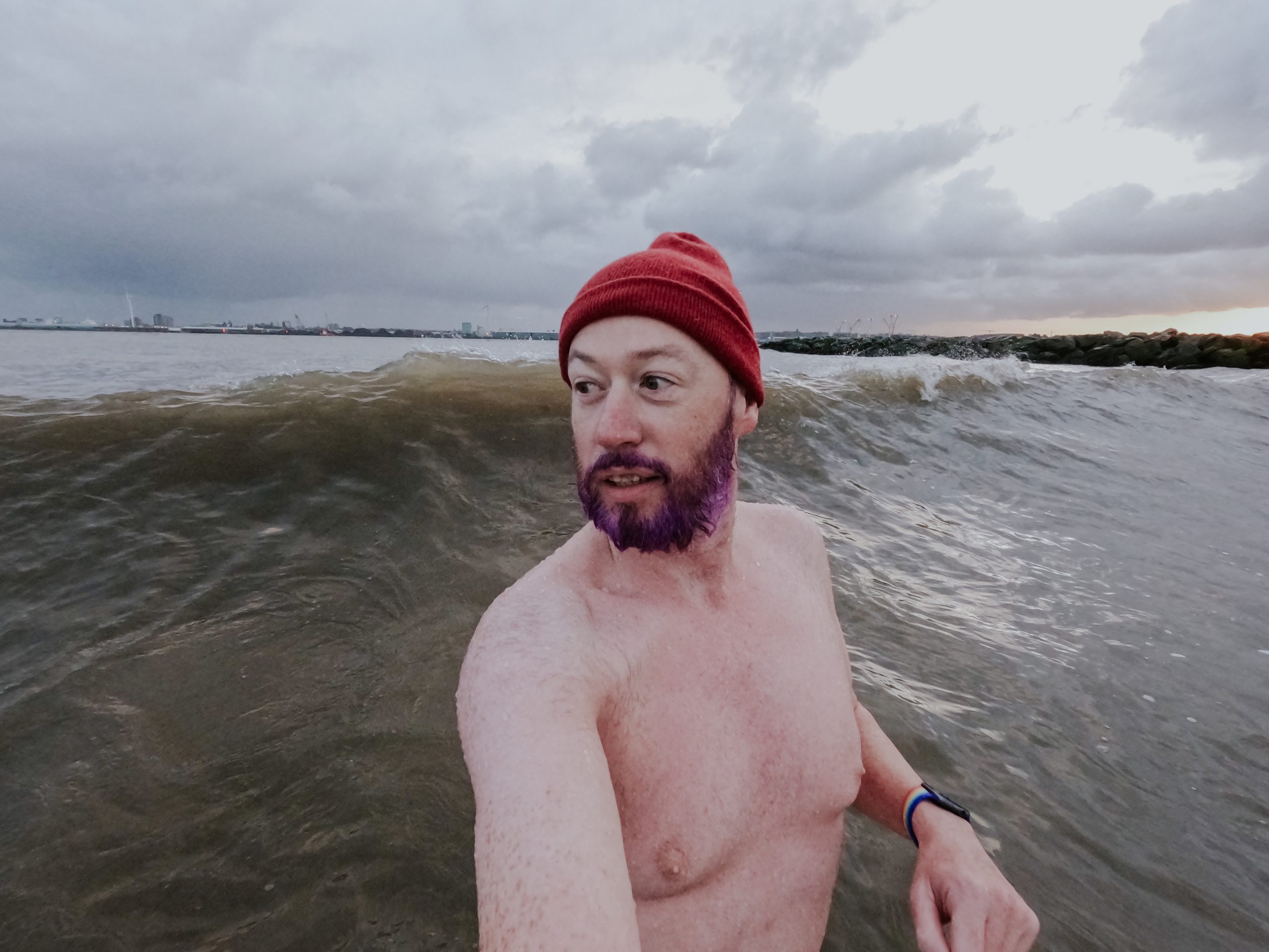 Non-binary person with a dark purple beard and a red beanie hat on glances back at a big wave closing in on them.