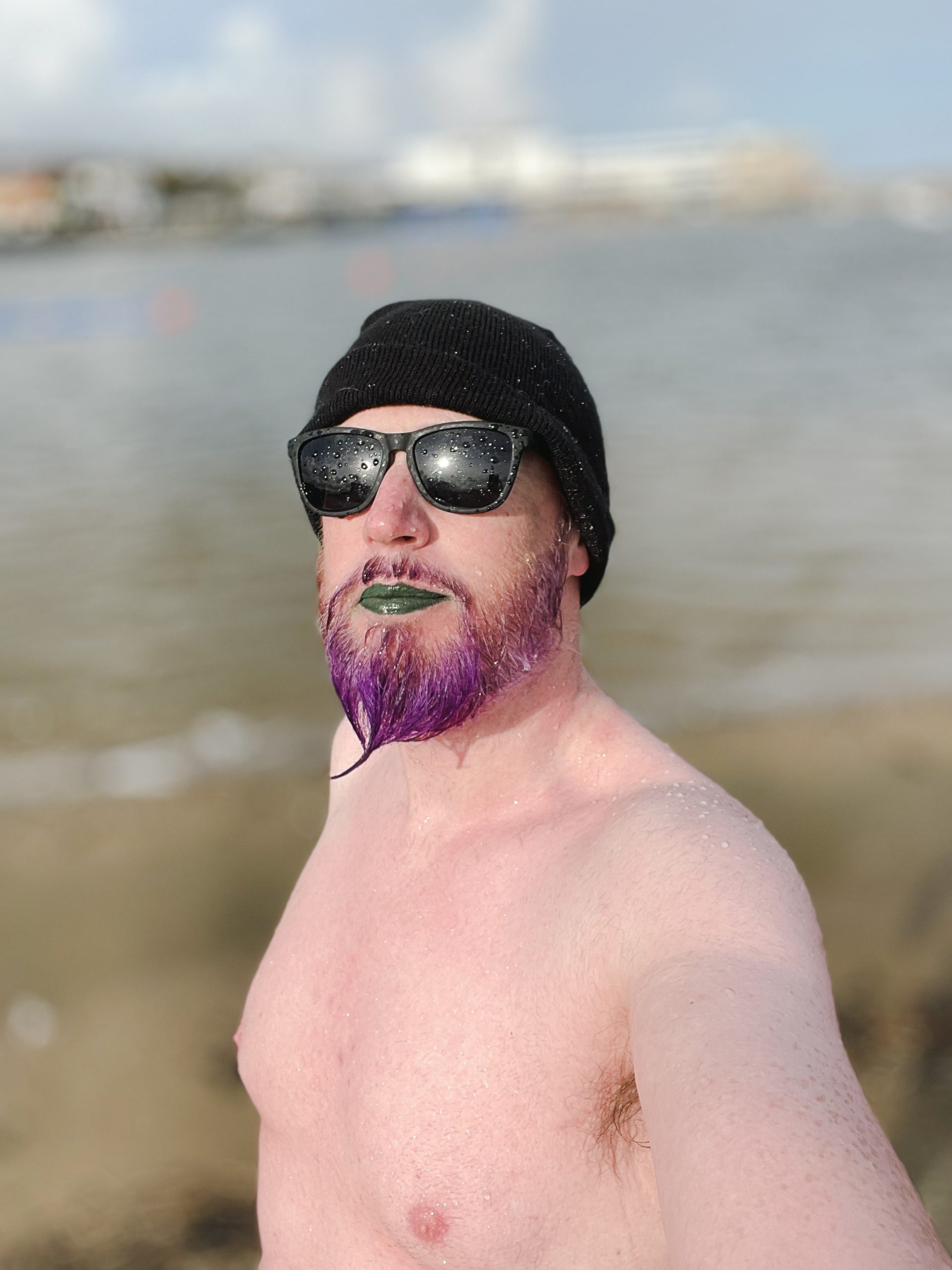 Non-binary person with a purple bead, green lipstick, black shades and a black beanie looks up to the sky after a swim.