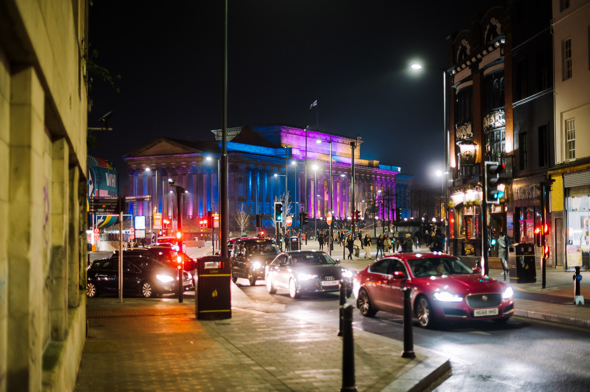 Walking down a street with busy traffic on. In the distance is St George's Hall which is lit up in the trans flag colours.