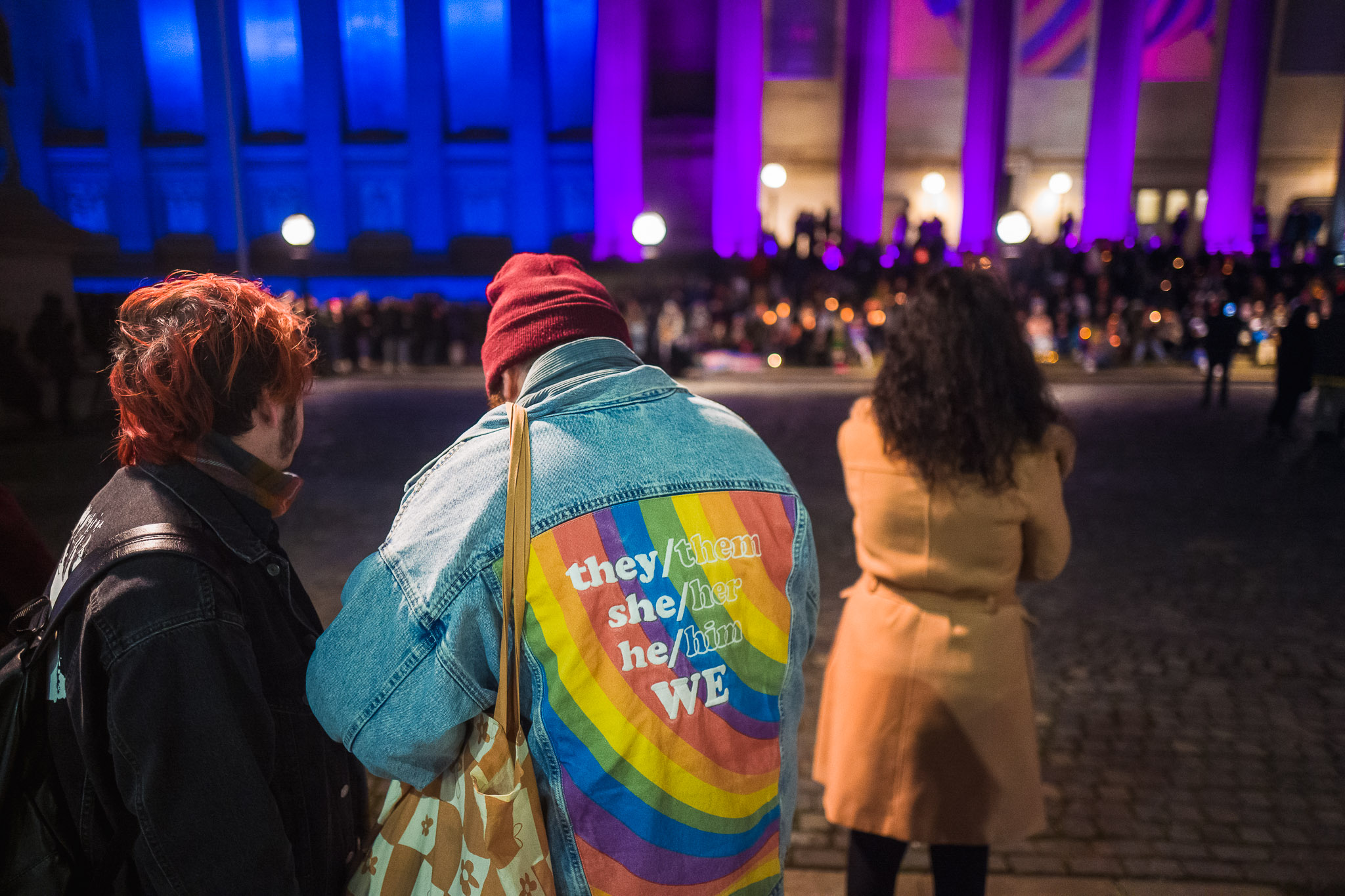 Standing on the plateau of St George's Hall someone is wearing a denim jacket with the message "They/them, she/her, he/him, we." on.