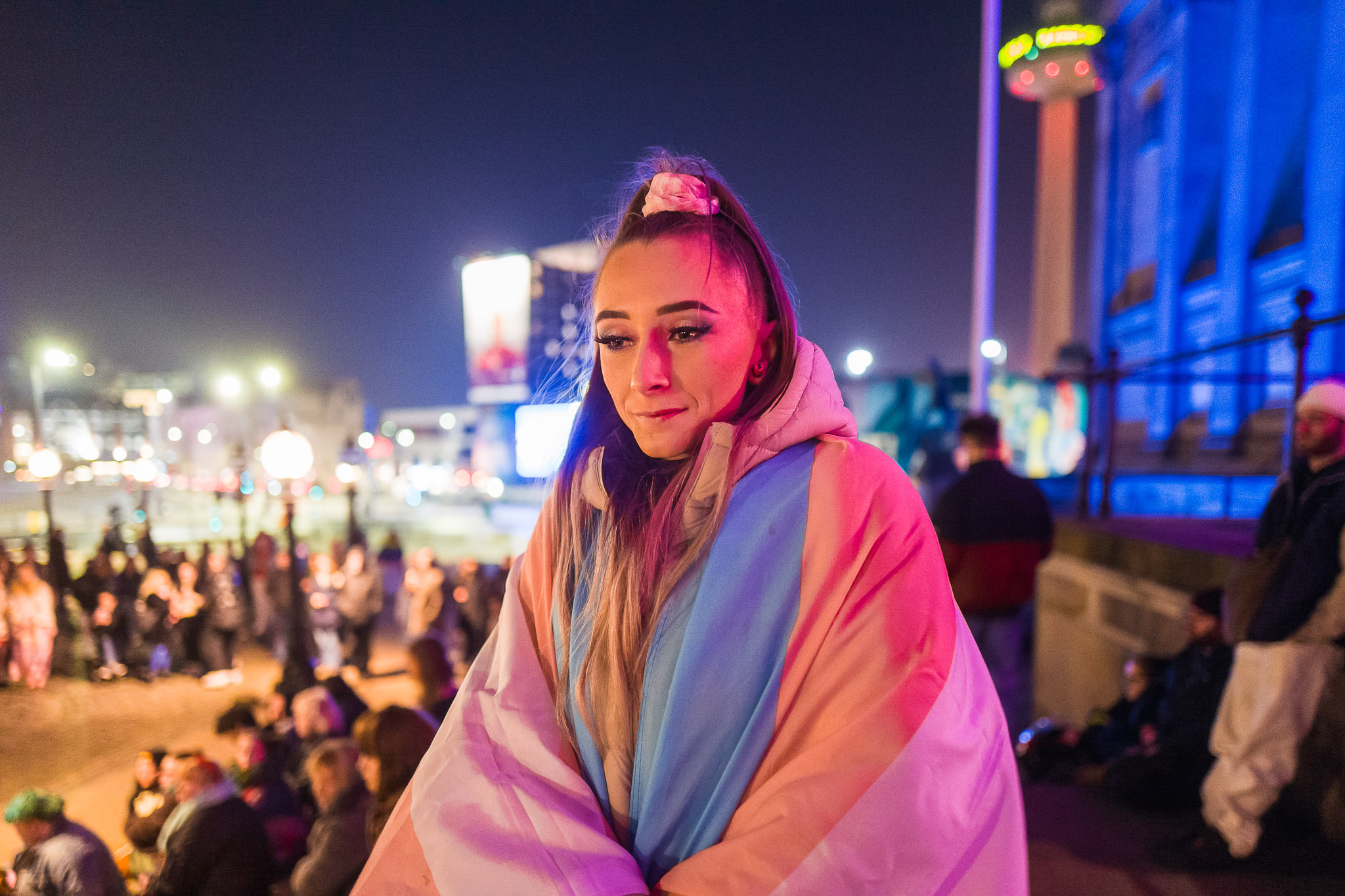 Portrait of a person wrapped in a trans flag on the steps of St George's Hall. St John's Beacon is in the background.