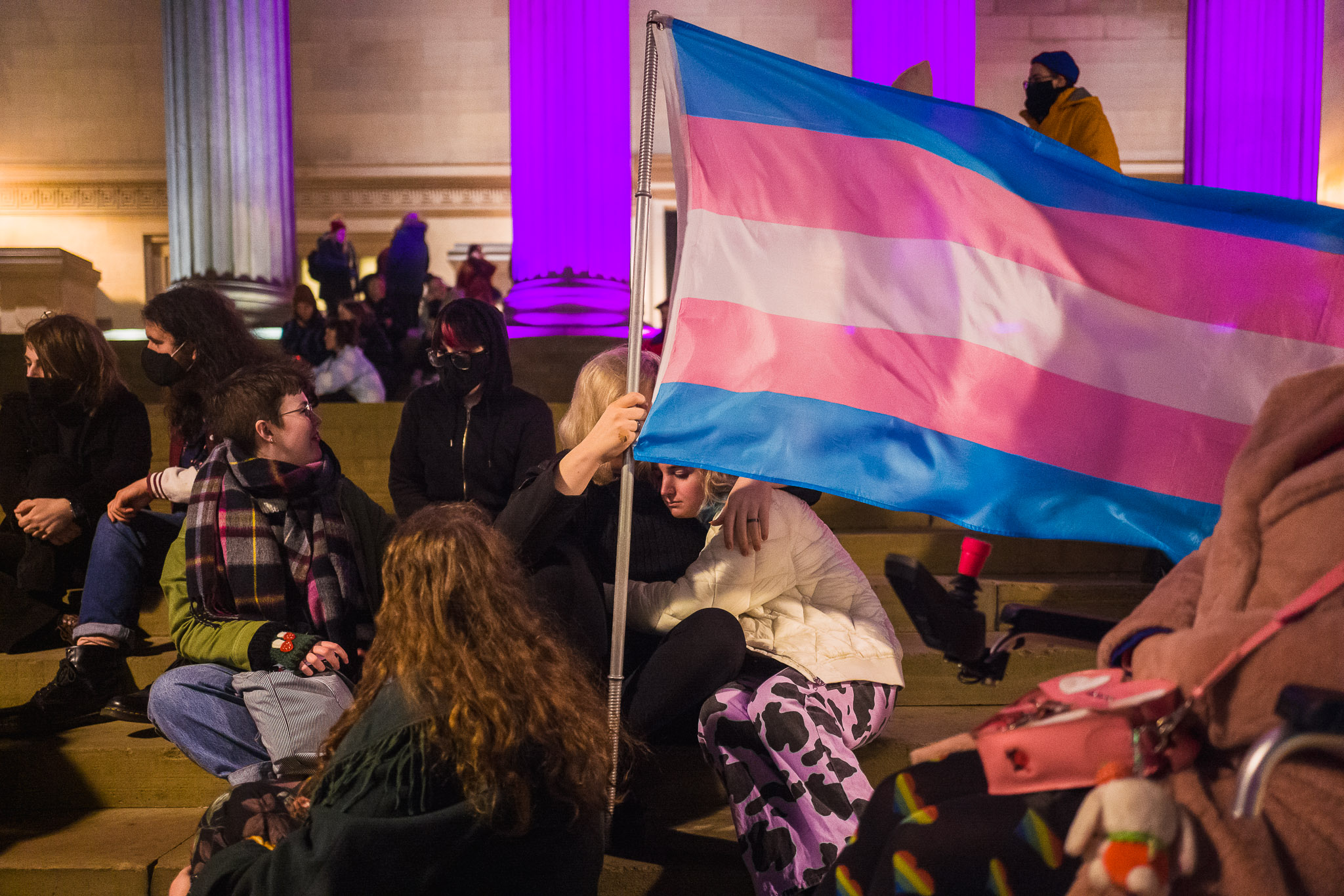 People sat on the steps of St George's Hall. Behind them the columns are pink. In the middle two people hug while one holds up a trans flag.