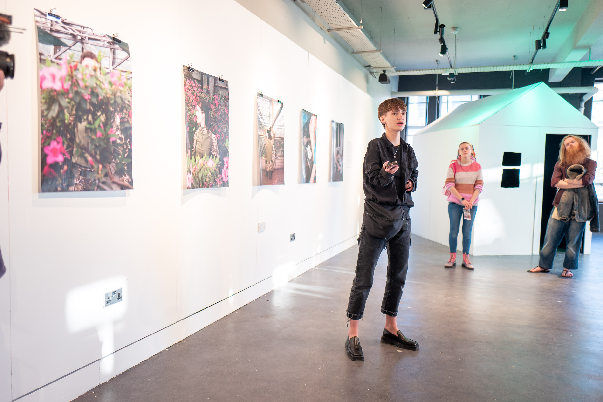A woman addresses an audience at a gallery launch. There are photos of pink flowers and a woman in military gear behind them.