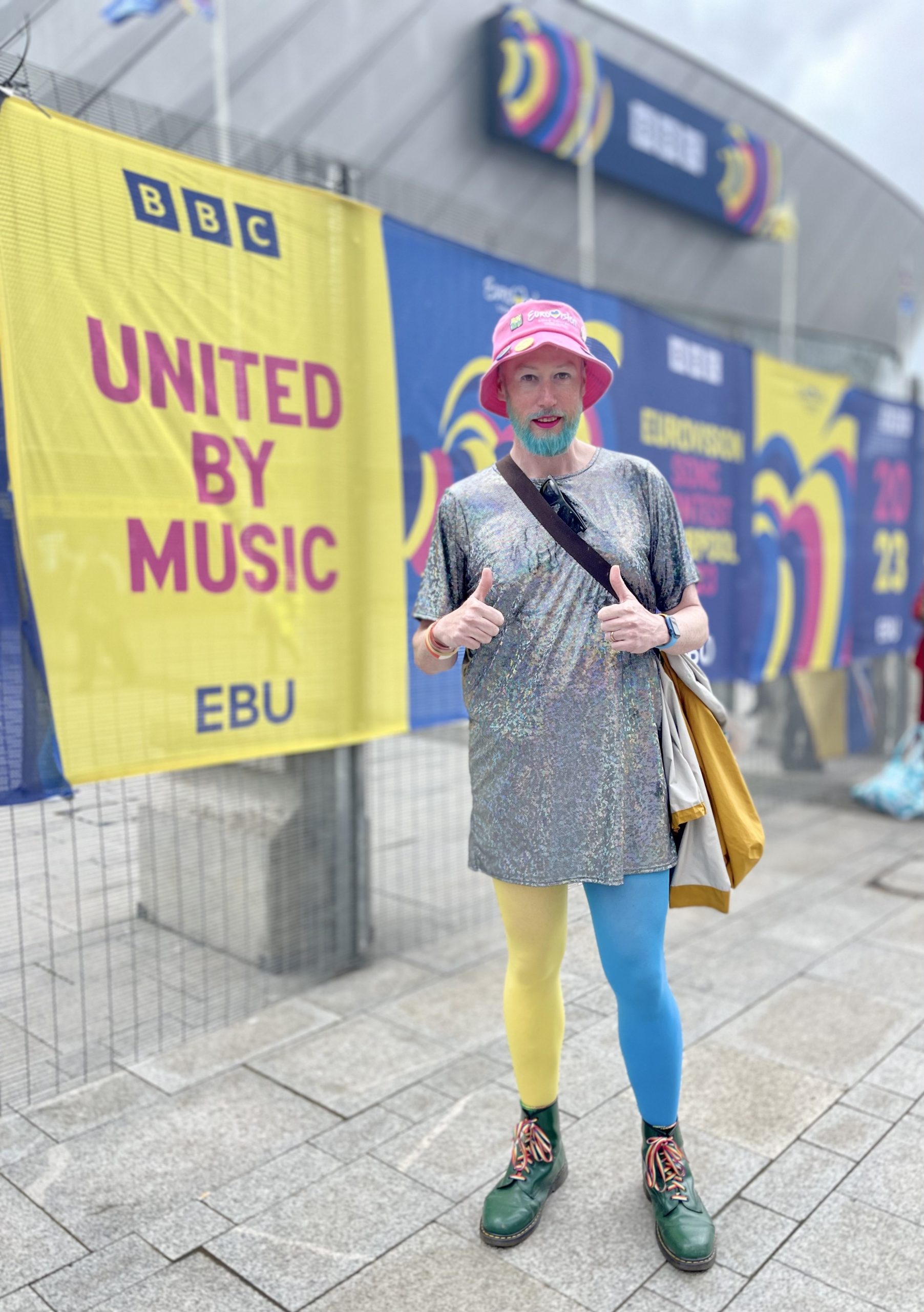 Non-binary person holding up two thumbs to the camera. They are wearing a sparkly t-shirt dress and blue and yellow tights, the colours of the Ukrainian flag. They are stood next to a sign saying "United by Music."