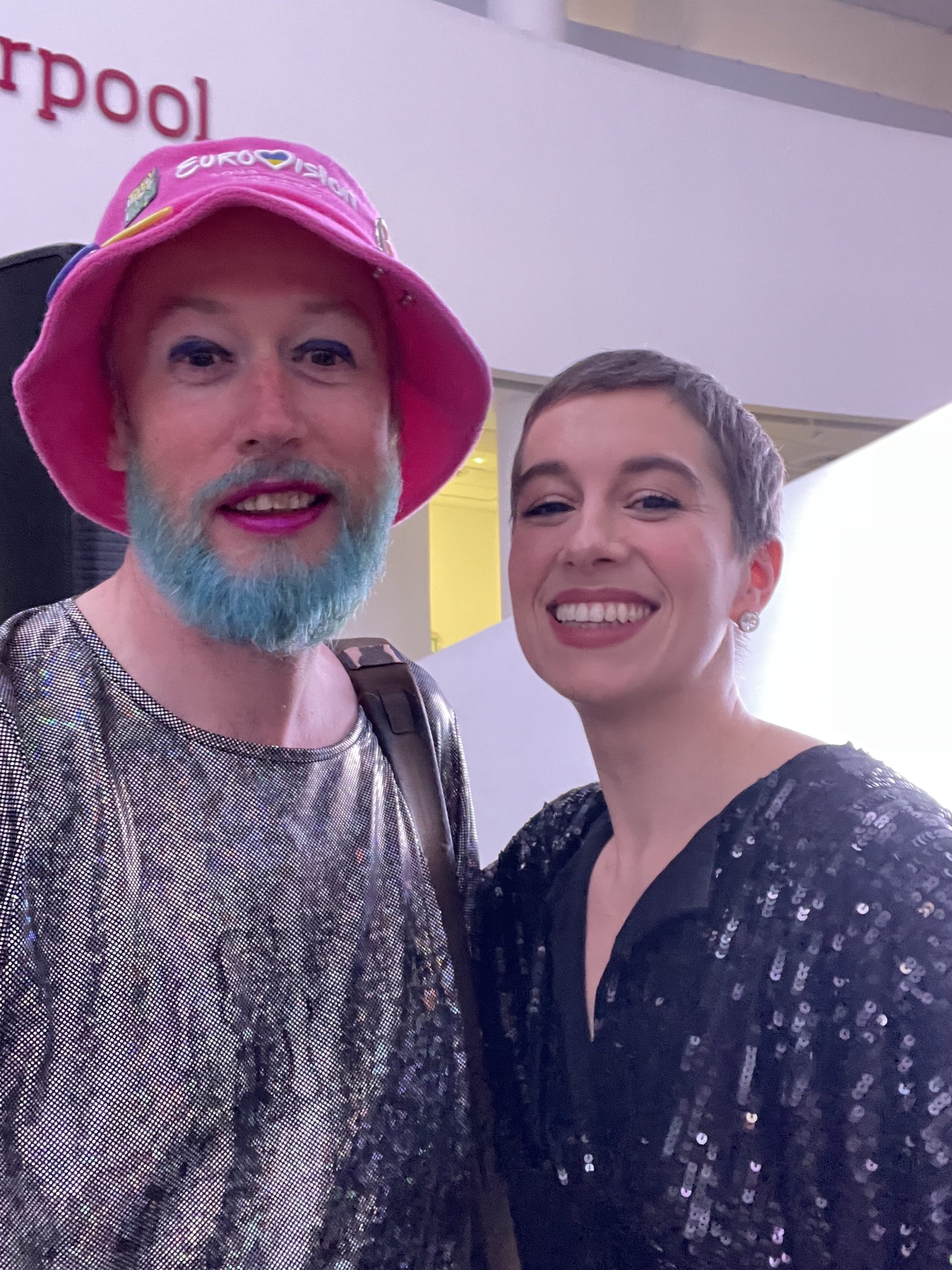 Non-binary person on the left in a sparkly dress, with blue beard and pink hat. Woman singer on the right in black sequin dress. Both smiling to camera.