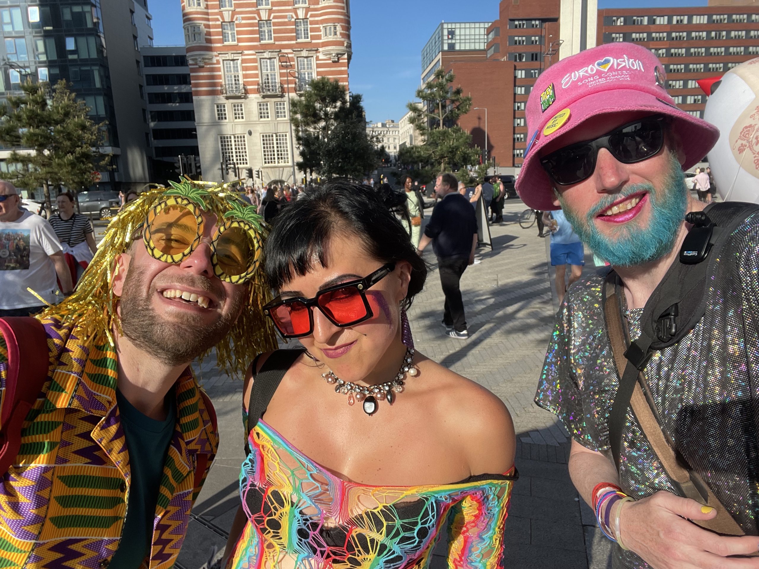 Non-binary person on the right in a sparkly dress, with blue beard and pink hat. Couple on the left in rainbow colours. Both smiling to camera.