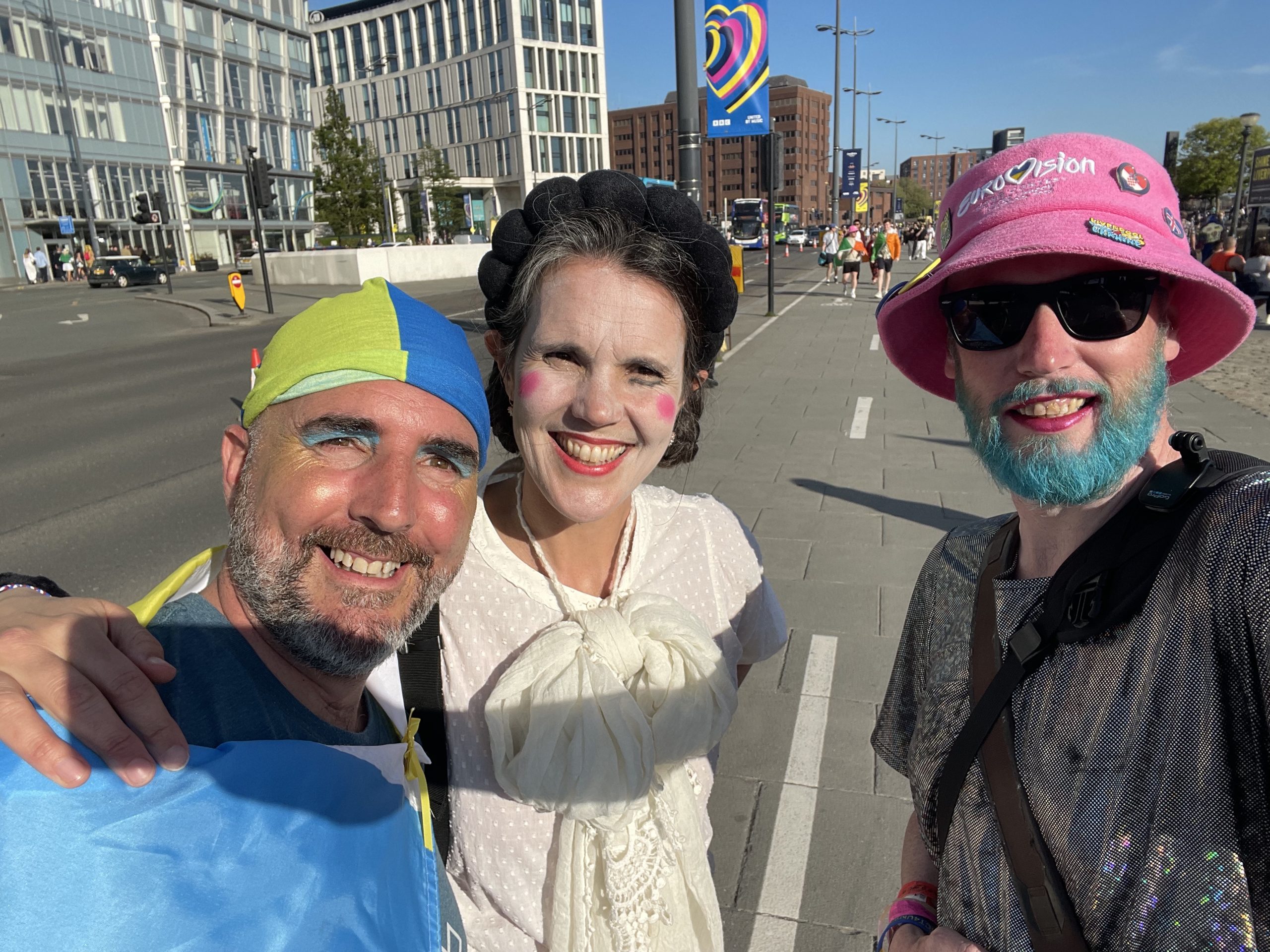 Non-binary person on the right in a sparkly dress, with blue beard and pink hat. Couple on the left in Ukrainian colours. Both smiling to camera.