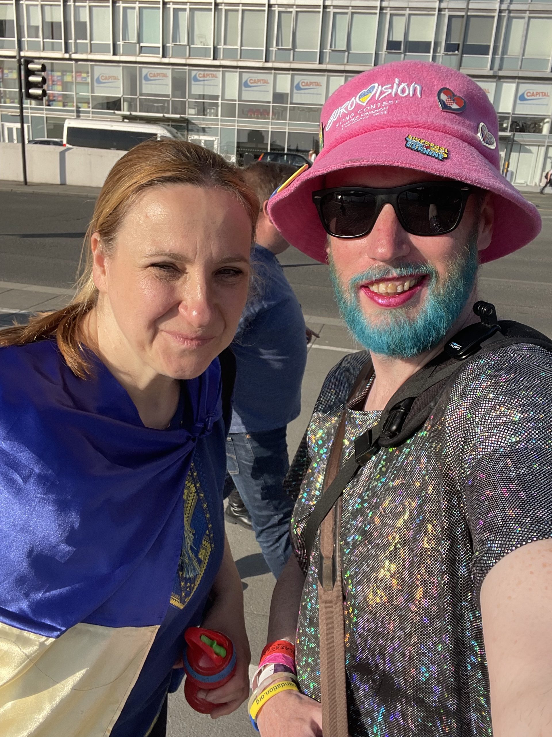 Non-binary person on the right in a sparkly dress, with blue beard and pink hat. Ukrainian woman on the left. Both smiling to camera.