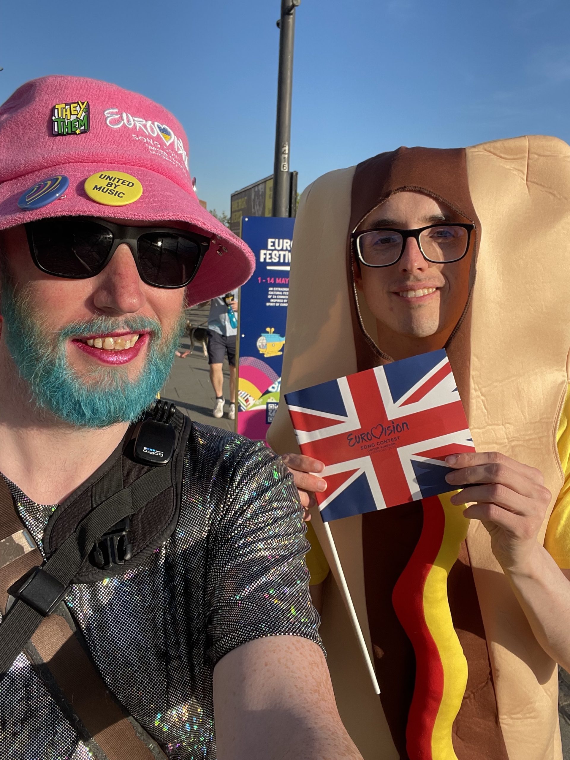 Non-binary person on the left in a sparkly dress, with blue beard and pink hat. Person dressed as hot dog with UK flag on the right. Both smiling to camera.